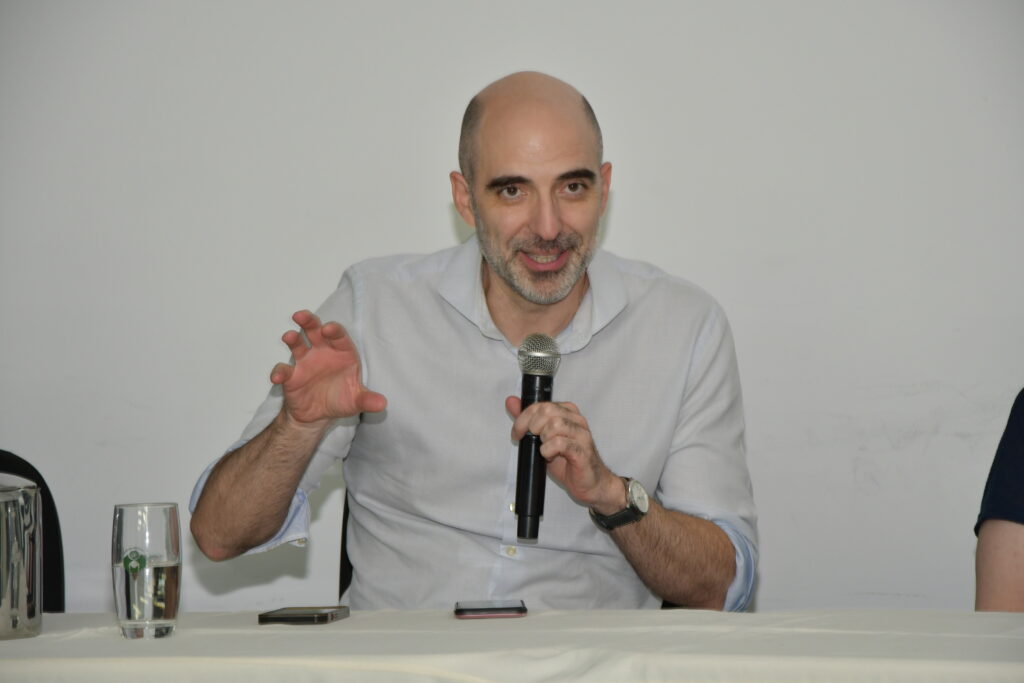 Um homem calvo e de barba grisalha, usando camisa social clara, fala ao microfone sentado à mesa de um evento. Ele gesticula com uma das mãos, enquanto segura o microfone com a outra. À sua frente há um copo com água, um celular e um jarro metálico.