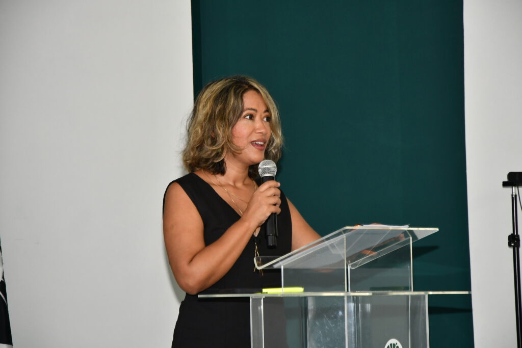 Uma mulher de cabelos loiros com mechas, usando vestido preto sem mangas, fala ao microfone posicionada atrás de um púlpito de acrílico. O fundo é branco e verde-escuro.