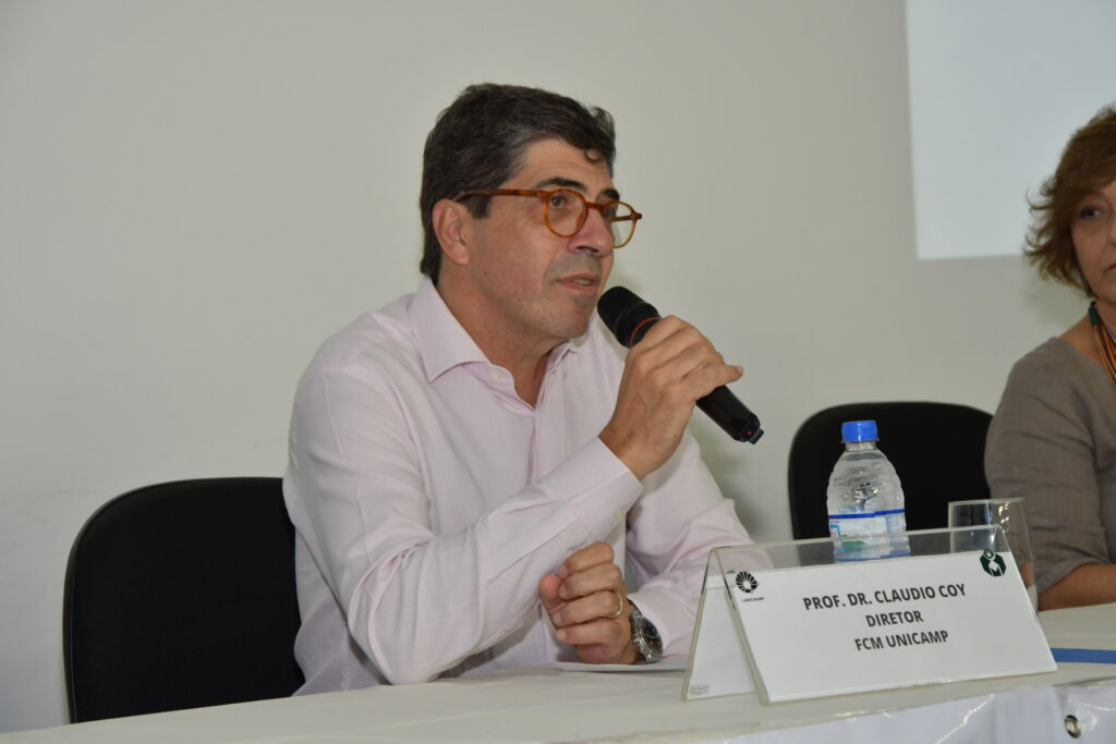 Um homem de óculos e camisa social rosa claro, identificado pela placa à sua frente como "Prof. Dr. Claudio Coy, Diretor FCM Unicamp", fala ao microfone em uma mesa de evento. Sobre a mesa há uma garrafa de água, um copo e uma mulher parcialmente visível ao fundo.