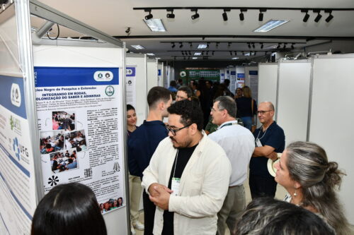 Pessoas observam e discutem pôsteres científicos expostos em painéis durante um evento acadêmico realizado em um espaço interno iluminado. Em primeiro plano, um homem de óculos e camisa clara conversa com outros participantes em frente a um pôster intitulado “Integrando em Rodas, Descolonização do Saber e Adinkras”, que contém textos, símbolos e fotos de atividades em grupo. Ao fundo, há mais pessoas circulando e interagindo entre os estandes.