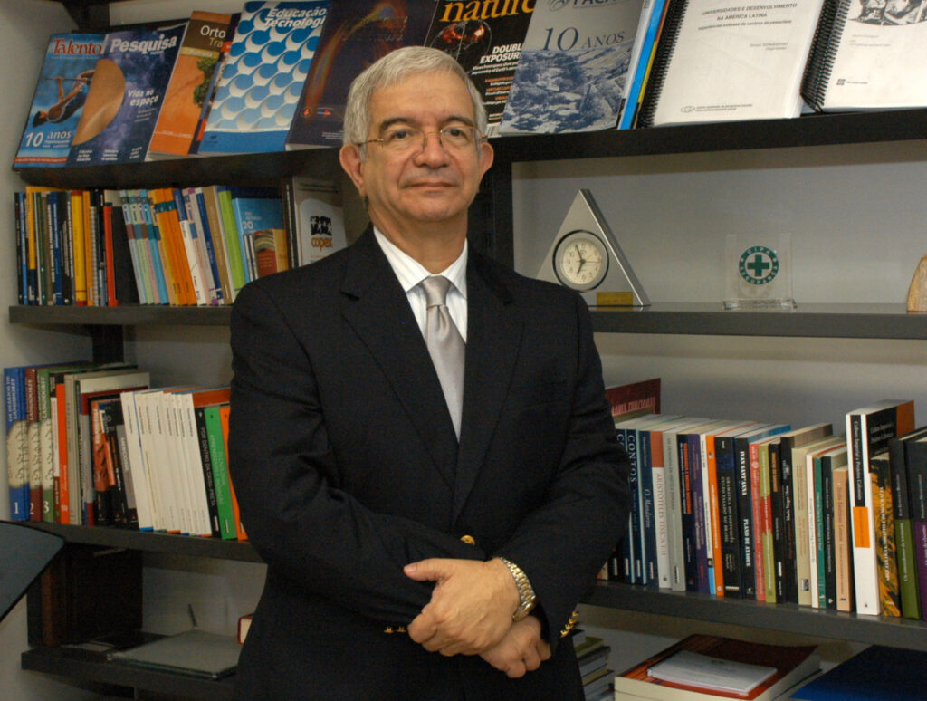 O professor Fernando costa é um homem de cabelos grisalhos usando óculos, terno escuro, camisa branca e gravata prata, em pé diante de estantes repletas de livros e revistas acadêmicas. Ele está com os braços cruzados, olhando para a câmera, em um ambiente que parece ser um escritório ou biblioteca