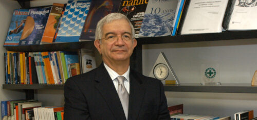 O professor Fernando costa é um homem de cabelos grisalhos usando óculos, terno escuro, camisa branca e gravata prata, em pé diante de estantes repletas de livros e revistas acadêmicas. Ele está com os braços cruzados, olhando para a câmera, em um ambiente que parece ser um escritório ou biblioteca