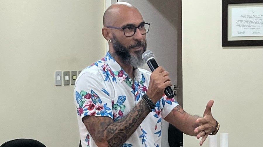 Homem com barba e óculos de grau, vestido com uma camisa floral colorida, fala ao microfone em um evento.