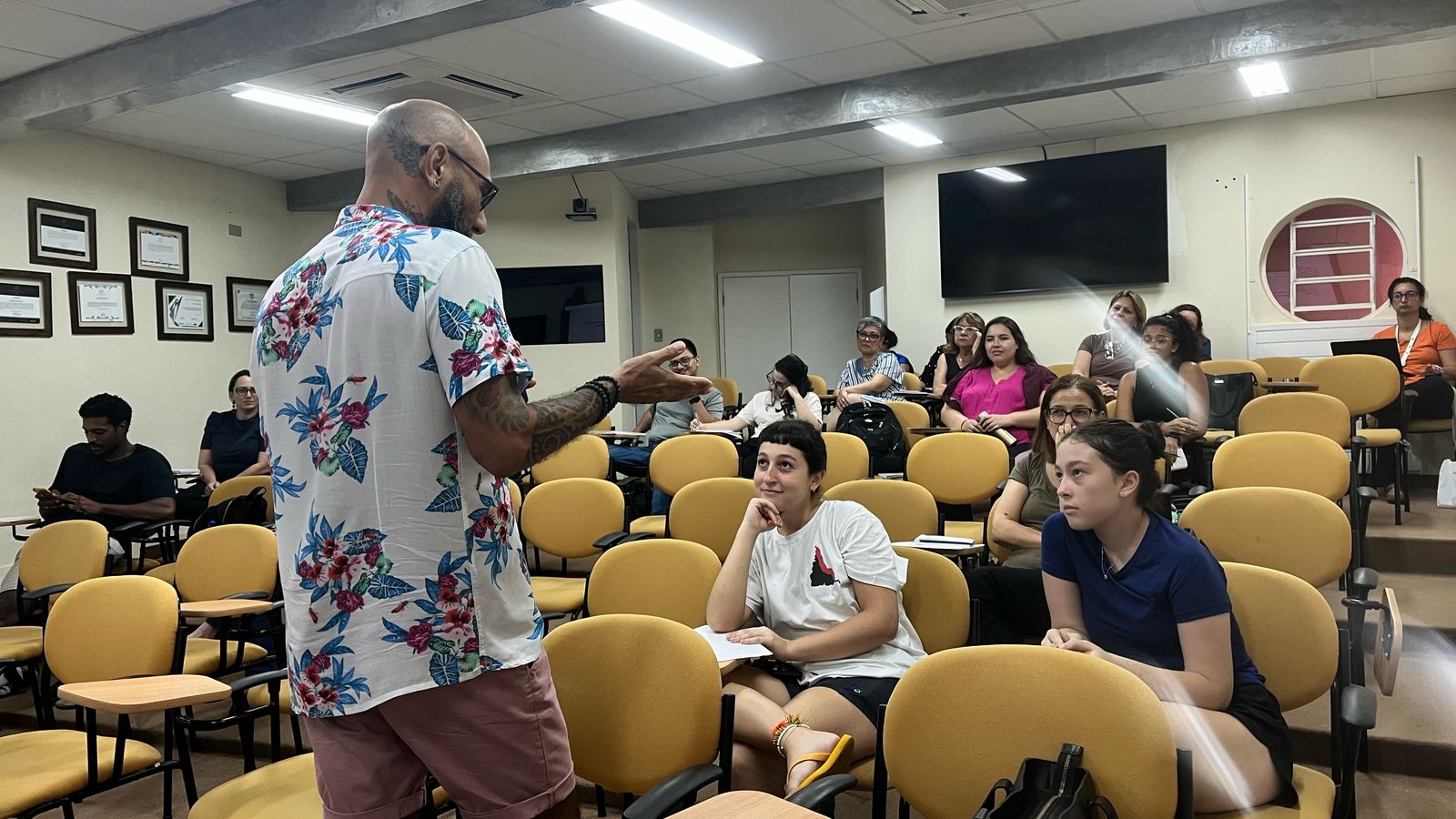 Pessoas participando de uma aula interativa, com um professor apresentando em um ambiente de aprendizado. Estudantes envolvidos e atentos.