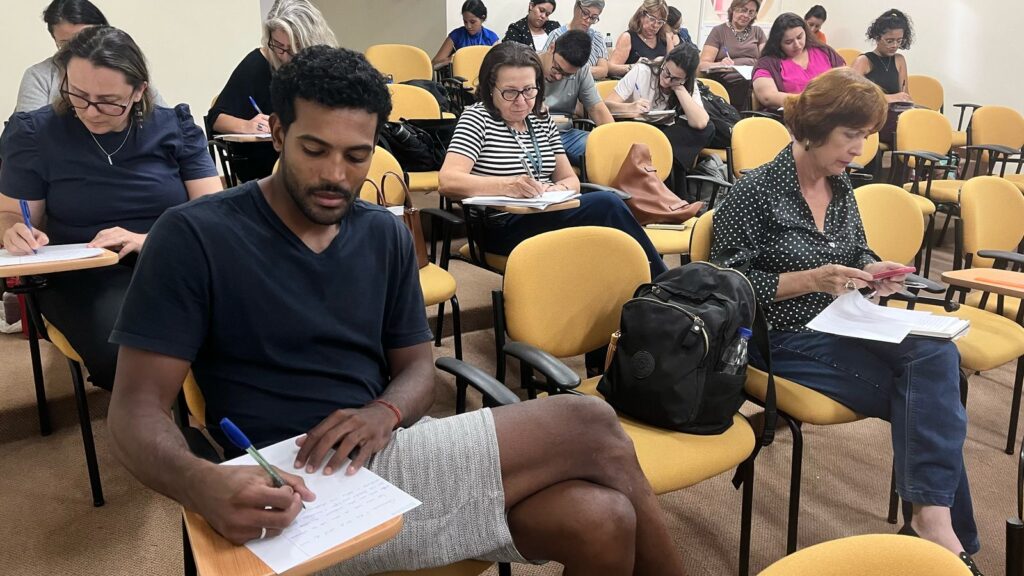Grupo de pessoas assistindo a uma aula e fazendo anotações em um ambiente educacional. Atenção e foco são visíveis em cada aluno.