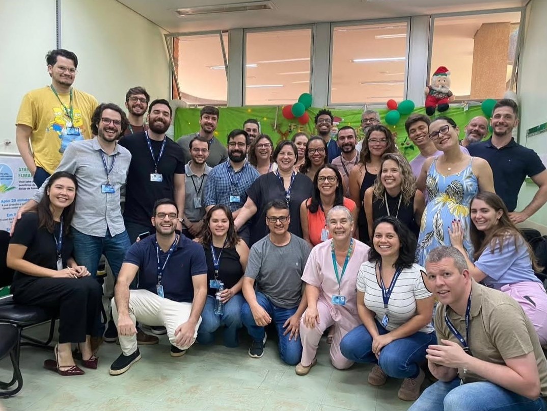 Grupo de aproximadamente 30 pessoas reunidas em uma sala, posando sorridentes para a foto. A maioria usa crachá e roupas informais. Ao fundo, há uma decoração verde com balões vermelhos e verdes e um boneco de Papai Noel, sugerindo um clima de confraternização de fim de ano. Algumas pessoas estão sentadas na frente, outras agachadas ou em pé, formando três fileiras.