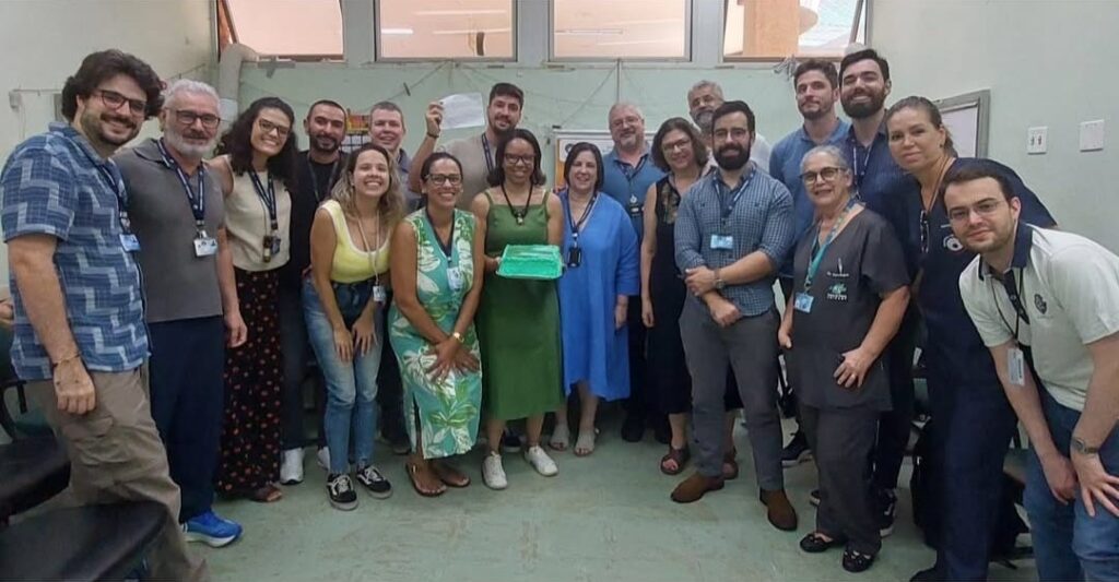 Grupo de cerca de 25 pessoas posando para foto em uma sala com janelas ao fundo. No centro da imagem, duas mulheres seguram uma travessa com bolo verde. O grupo está sorridente e aparenta celebrar uma ocasião especial. A maioria usa crachá e roupas casuais ou de trabalho. A disposição é mais compacta, com pessoas lado a lado em pé.