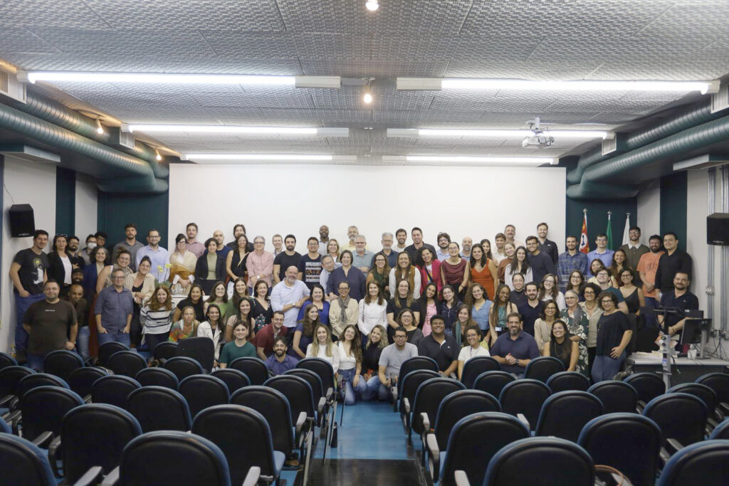 Grande grupo de pessoas reunido em um auditório para uma foto coletiva. Estão todos sorrindo, com fileiras de cadeiras vazias à frente e uma tela de projeção ao fundo.
