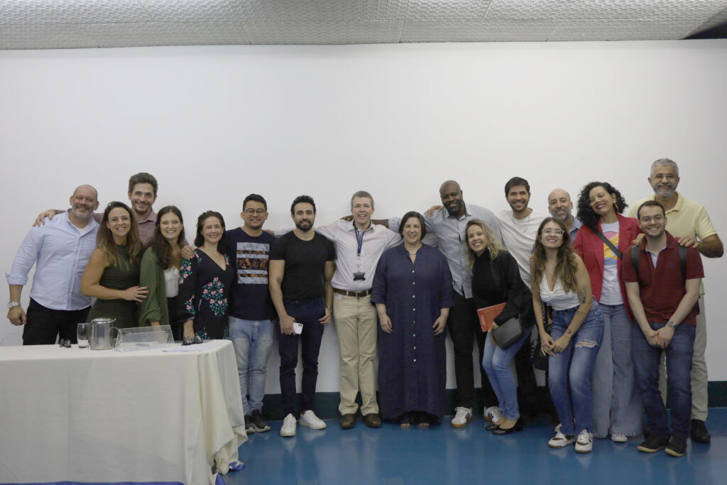 Grupo menor de pessoas posando sorridentes em frente a uma parede branca, com uma mesa coberta por uma toalha clara à esquerda.