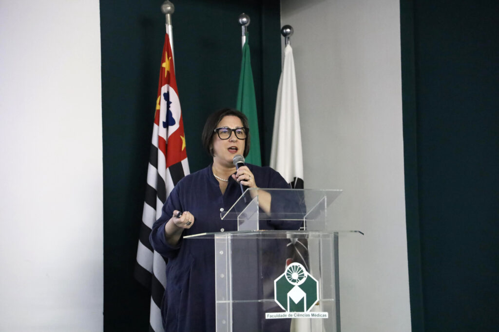 Mulher falando ao microfone em um púlpito transparente com o logotipo da Faculdade de Ciências Médicas. Ao fundo, bandeiras do Brasil, do estado de São Paulo e da instituição.