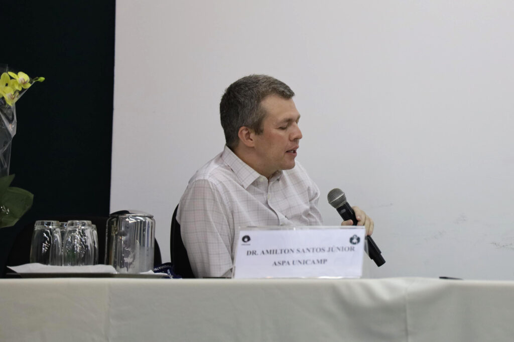 Um homem de cabelos curtos e grisalhos, vestindo uma camisa clara xadrez, está sentado em uma mesa de conferência segurando um microfone. À sua frente há uma placa com seu nome: "Dr. Amilton Santos Júnior - ASPA UNICAMP", além de copos e uma jarra de água.