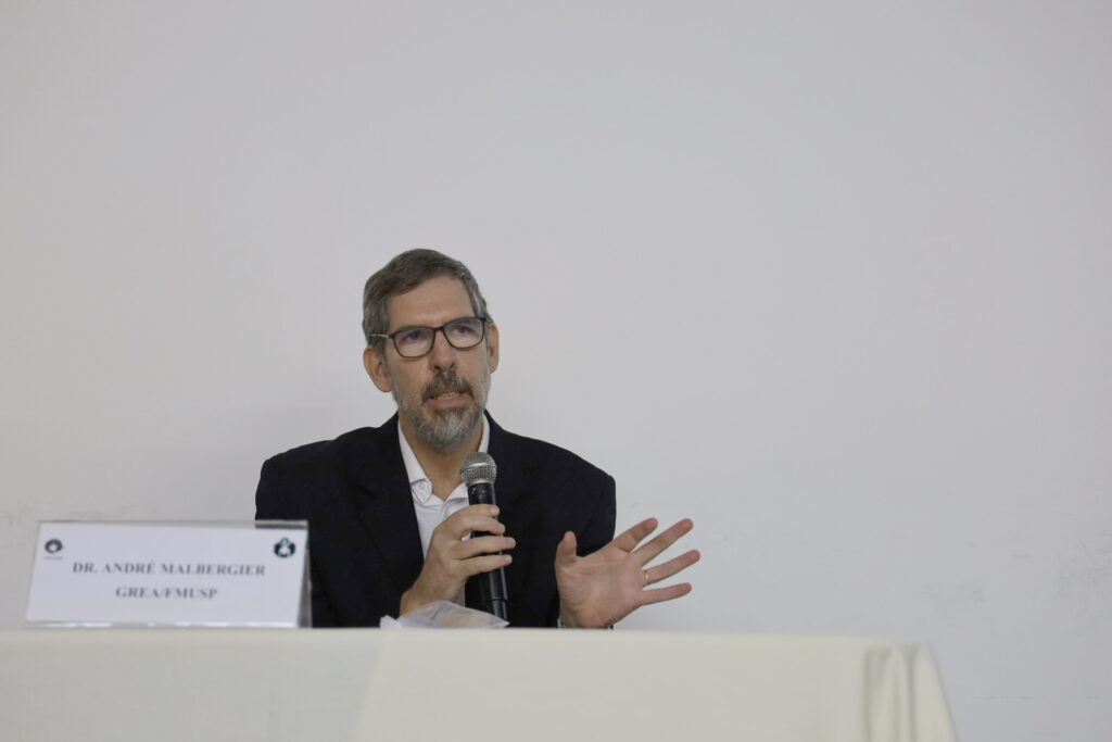 Um homem com barba grisalha e óculos, usando um paletó escuro, está falando enquanto segura um microfone com uma das mãos e gesticula com a outra. Na frente dele há uma placa com seu nome: "Dr. André Malbergier - GREA/FMUSP"