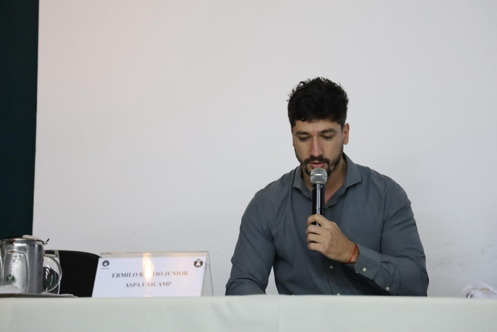 Um homem jovem, com cabelo escuro e barba, usando uma camisa social cinza, está sentado e lendo algo enquanto segura um microfone próximo à boca. À sua frente há uma placa com o nome: "Ermilo Benito Junior - ASPA UNICAMP