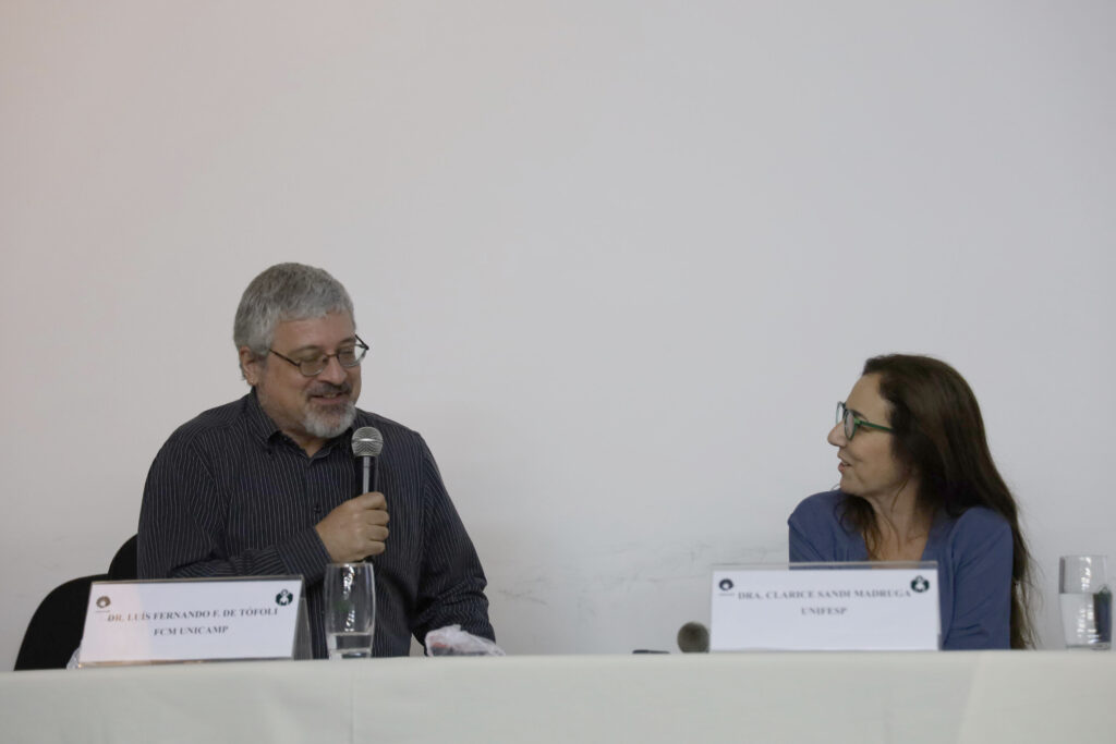 Dois palestrantes estão sentados lado a lado. À esquerda, um homem grisalho com barba e óculos, segurando um microfone, está sorrindo. À direita, uma mulher de cabelos longos castanhos e óculos verdes o observa com um leve sorriso. As placas de identificação à frente deles leem: "Dr. Luís Fernando E. de Tófoli - FCM UNICAMP" e "Dra. Clarice Sandi Madruga - UNIFESP"