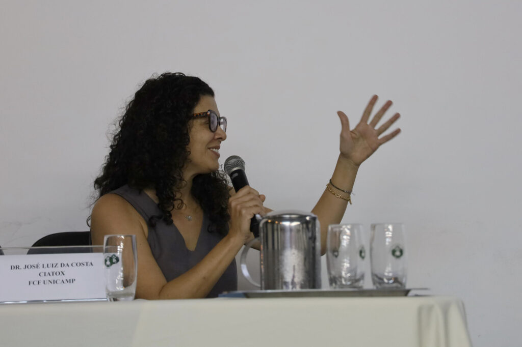 Uma mulher de cabelos cacheados e óculos fala ao microfone enquanto gesticula com a mão esquerda. Ela está sentada à mesa de um evento, ao lado de copos de vidro e uma jarra de metal. Uma placa identificadora ao lado tem o nome "Dr. José Luiz da Costa - CIATOX - FCF UNICAMP".