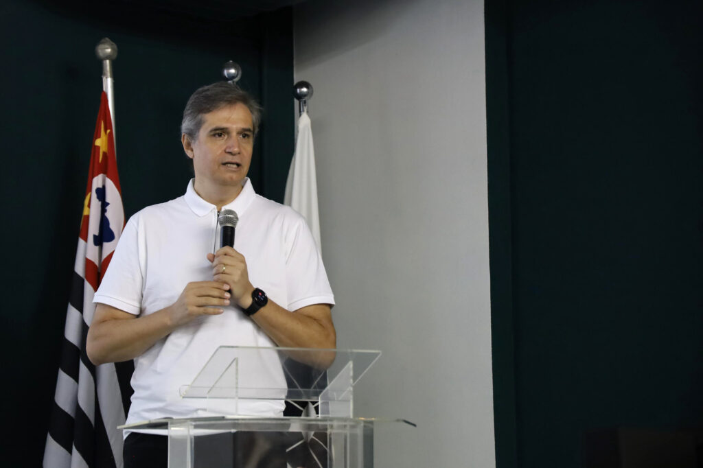 Um homem de cabelo grisalho, vestindo camisa polo branca, fala ao microfone em pé atrás de um púlpito de acrílico. Atrás dele estão as bandeiras do estado de São Paulo e do Brasil.