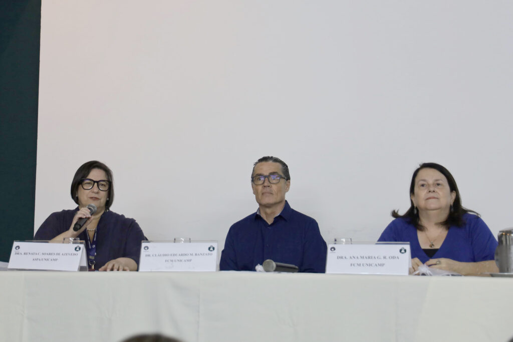 Três pessoas estão sentadas à mesa durante um evento. À esquerda, uma mulher segura um microfone e fala, enquanto duas outras pessoas, um homem ao centro e uma mulher à direita, observam. Placas identificam os participantes como membros da UNICAMP.