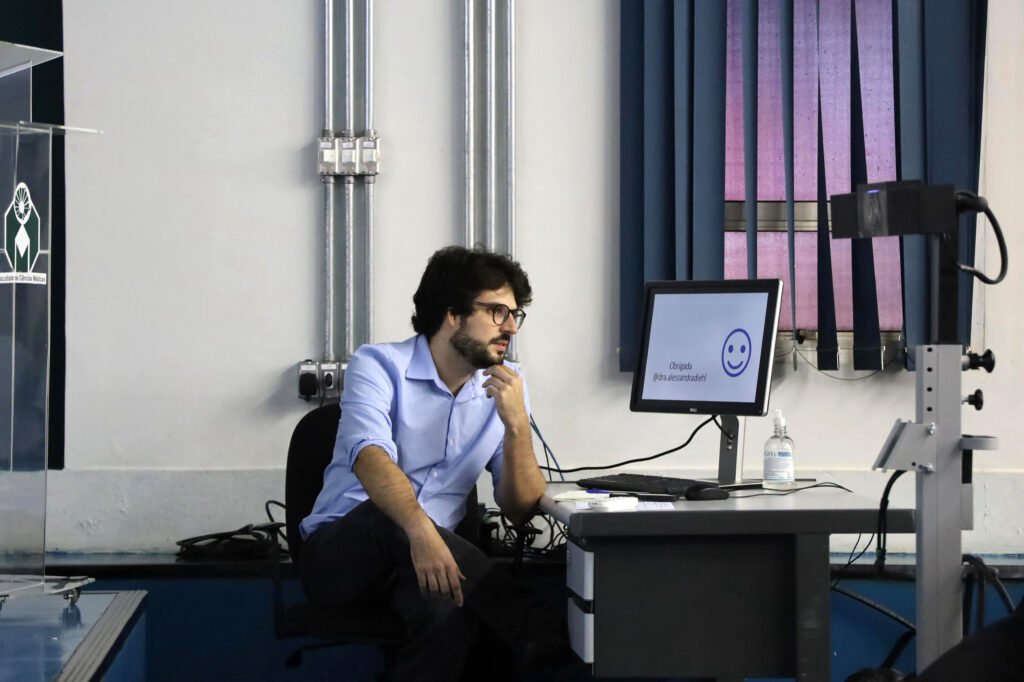 Um homem de barba e óculos está sentado à frente de um computador, com a mão no queixo em pose pensativa. Na tela do monitor aparece um emoji sorridente e a mensagem “Obrigada @dra.alessandrafel”. Há uma garrafa de álcool em gel ao lado do computador.