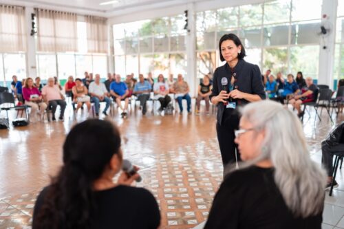A professora Laura Moryiama, em pé, segura um microfone enquanto interage com duas mulheres sentadas, em um grande salão iluminado. Ao fundo, um grupo de pessoas está sentado em círculo, participando de uma reunião ou roda de conversa. O ambiente tem piso de madeira e amplas janelas que permitem a entrada de luz natural.