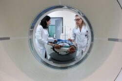 Vista de dentro de um aparelho de tomografia computadorizada, mostrando duas profissionais da saúde vestidas com jaleco branco preparando um paciente de pele escura, que está deitado na maca coberto com um cobertor xadrez. A cena ocorre em um ambiente hospitalar com equipamentos médicos ao fundo.