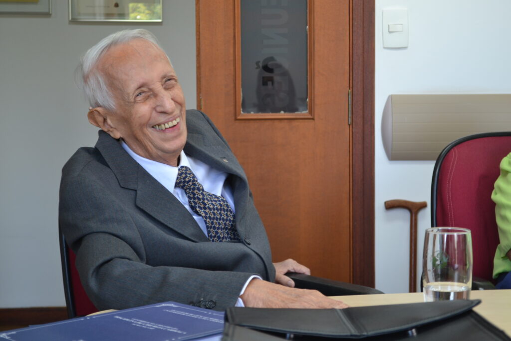 O médico cirurgião Antônio Benedicto Prado Fortuna está sentado com o braço direito apoiado na mesa. Ele usa terno e gravata e sorri para a foto. Ao fundo da imagem é possível notar a sua bengala apoiada na parede,