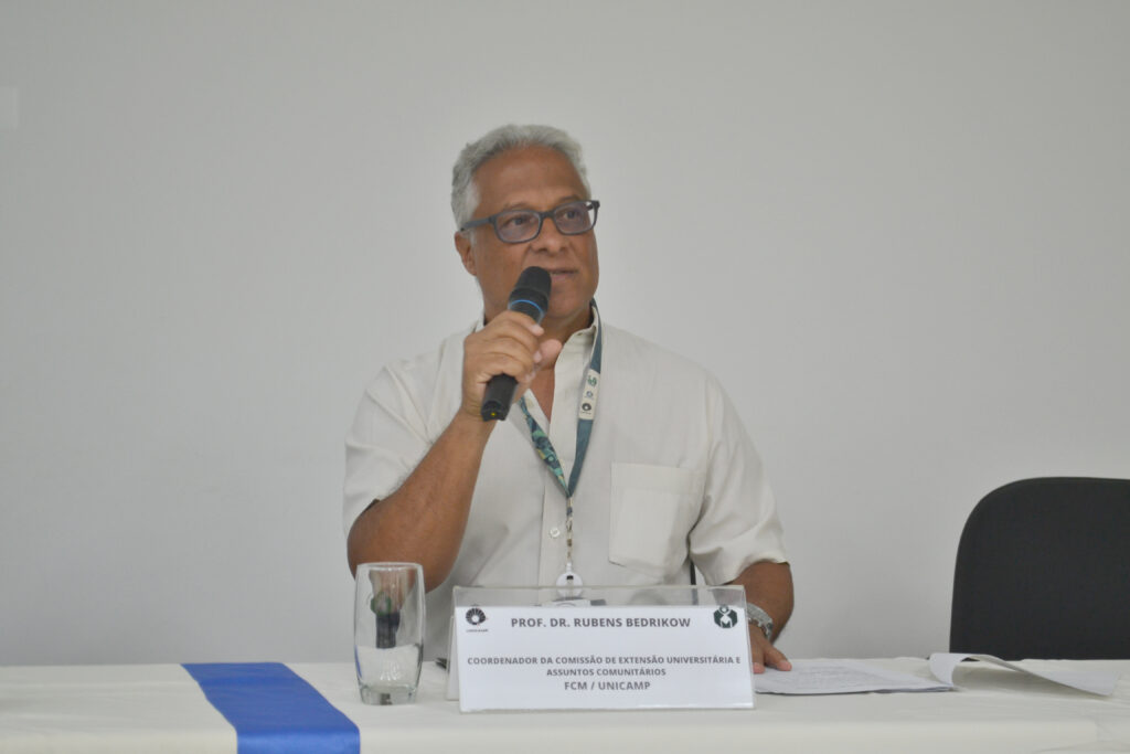 Professor Dr. Rubens Bedrikow durante uma apresentação, coordenando a Comissão de Extensão Universitária. Ele está segurando um microfone e falando para a audiência.