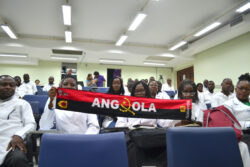 Pessoas em um auditório segurando uma bandeira de Angola, demonstrando orgulho e identidade cultural. Ambiente acadêmico com participação ativa dos estudantes.