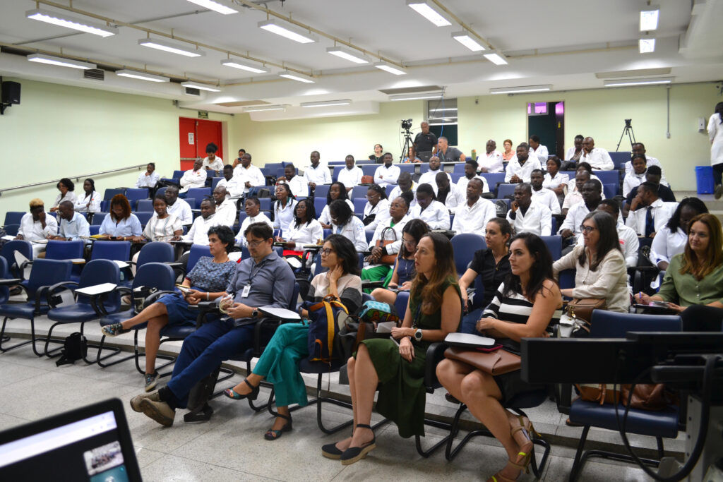 Imagem de um auditório lotado com participantes em polos brancos, prestando atenção em uma apresentação, destacando a interação acadêmica.