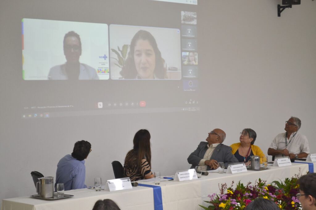 Imagem de uma videoconferência com vários participantes visíveis em uma mesa. Pessoas assistindo a um evento com apresentações online, com destaque para dois palestrantes no telão.
