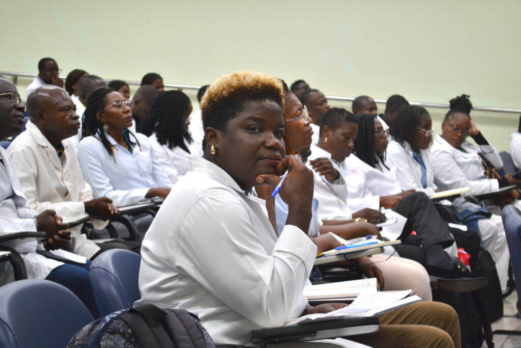 Imagem de um grupo de estudantes de medicina, em sala de aula, atentos durante uma aula, com anotações e em trajes brancos.