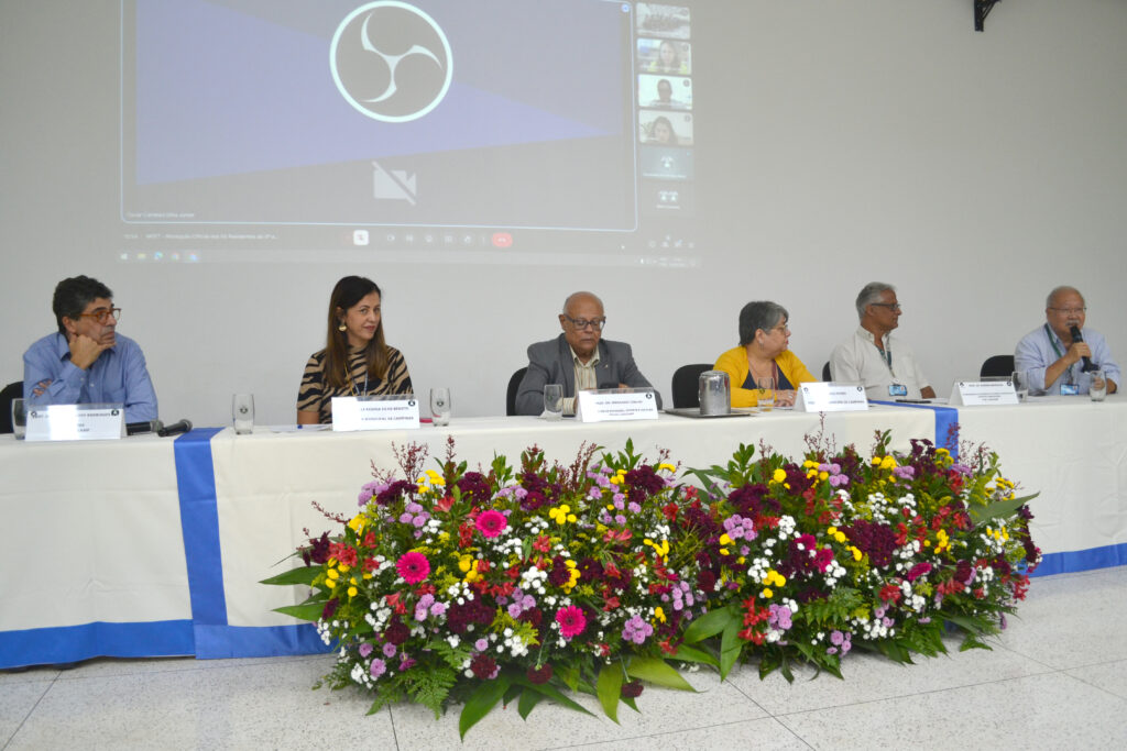 Mesa de debate com cinco pessoas sentadas, apresentando um evento, com flores coloridas na frente e uma tela ao fundo com logotipo do software de streaming.