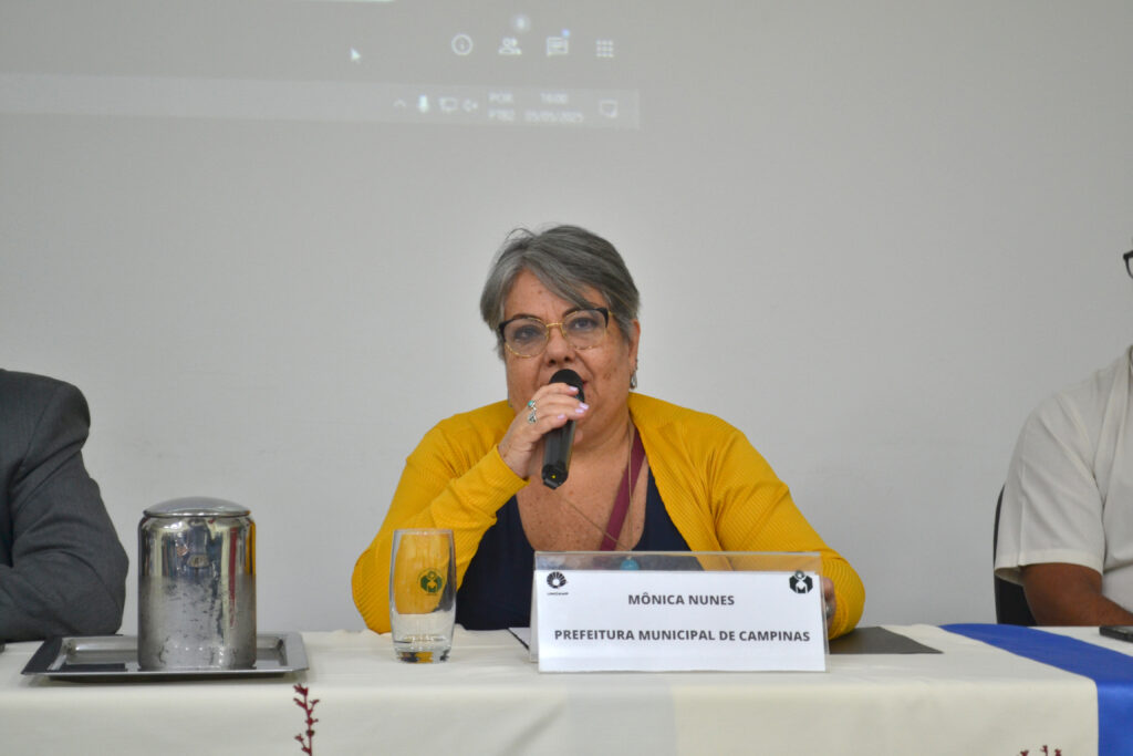 Mônica Nunes em evento da Prefeitura Municipal de Campinas. Ela fala ao microfone, representando a administração local com comprometimento e dedicação.