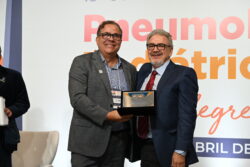 O professor José Dirceu Ribeiro, à direita, usa terno azul-marinho, camisa azul clara e gravata listrada em vermelho e azul. Ele sorri enquanto segura uma placa de homenagem ao lado de outro homem, que veste terno cinza e camisa azul. Ambos estão no palco do 18º Congresso Brasileiro de Pneumologia Pediátrica, com fundo branco e parte do letreiro do evento visível.