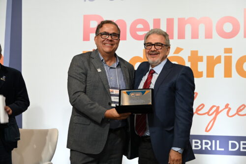 O professor José Dirceu Ribeiro, à direita, usa terno azul-marinho, camisa azul clara e gravata listrada em vermelho e azul. Ele sorri enquanto segura uma placa de homenagem ao lado de outro homem, que veste terno cinza e camisa azul. Ambos estão no palco do 18º Congresso Brasileiro de Pneumologia Pediátrica, com fundo branco e parte do letreiro do evento visível.