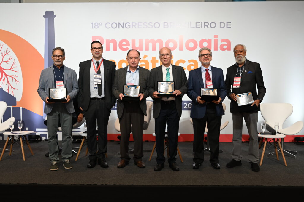 Seis homens estão perfilados no palco do 18º Congresso Brasileiro de Pneumologia Pediátrica, todos segurando placas de homenagem. Atrás deles, o painel do congresso traz ilustrações e o nome do evento. Os homenageados vestem trajes formais e posam para a foto em frente a cadeiras brancas e copos de água sobre mesas laterais.
