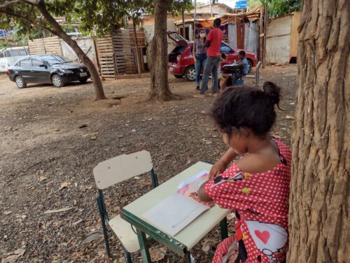 Menina com vestido vermelho de bolinhas brancas e estampa de personagem infantil sentada ao ar livre em uma pequena carteira escolar, próxima a uma árvore, concentrada em um livro ilustrado. Ao fundo, outras pessoas estão reunidas ao lado de um carro vermelho com o porta-malas aberto, em uma área com chão de terra e barracos construídos com madeira e telhas metálicas.