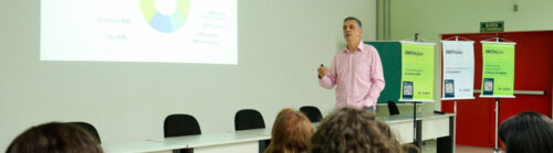 Um homem, de camisa rosa clara, está em pé ao lado de uma mesa em uma sala de aula ou auditório, apresentando uma palestra. Ele segura um controle remoto e fala para um grupo de pessoas sentadas, que aparecem de costas. Na parede à esquerda, há um projetor exibindo um gráfico de setores com dados de diferentes áreas, como “Automotivo”, “Agro”, “Saúde Humana e Animal”, entre outras. À direita, há três banners verticais com o título "INOVAÇÃO" e subtítulos como “Comunicação de invenção”, “Empreendedorismo e Incubadora” e “Defesas de dissertações e teses em sigilo”. Ao fundo, uma porta vermelha com uma placa verde de “SAÍDA DE EMERGÊNCIA”.