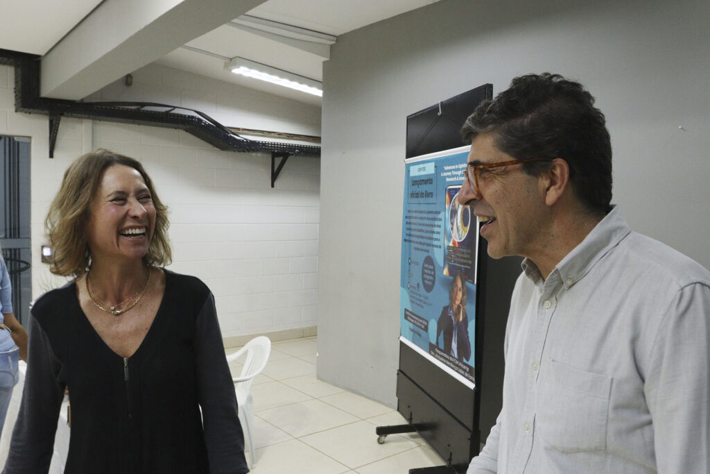 Mulher e homem sorrindo enquanto conversam em ambiente interno durante um evento. Ao fundo, um cartaz anuncia o lançamento oficial de um livro na área da oftalmologia. A atmosfera é descontraída e amigável, sugerindo confraternização em ambiente institucional.