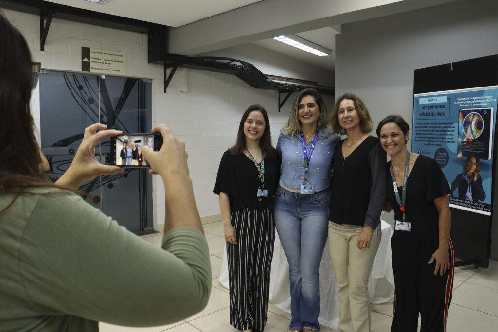 Grupo de quatro mulheres posando para uma foto, sendo fotografadas por uma quinta mulher com celular. Ao fundo, há uma porta de vidro com o nome “Laboratório de Citogenômica – GENOCa” e um cartaz anunciando o lançamento de um livro. As mulheres estão sorrindo e usam crachás, sugerindo um ambiente institucional.