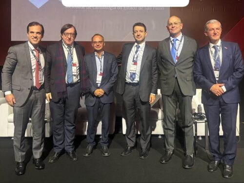 Seis homens posam juntos em pé sobre um palco durante um evento profissional. Todos vestem trajes sociais (terno e gravata) e usam crachás de identificação pendurados no pescoço. Ao fundo, vê-se parte de um telão com inscrições, parcialmente encobertas, onde se lê “Sociedade de Cardiologia do Estado de São Paulo”. O grupo está disposto lado a lado, sorrindo para a foto, e há poltronas brancas atrás deles, compondo o ambiente do auditório ou congresso.