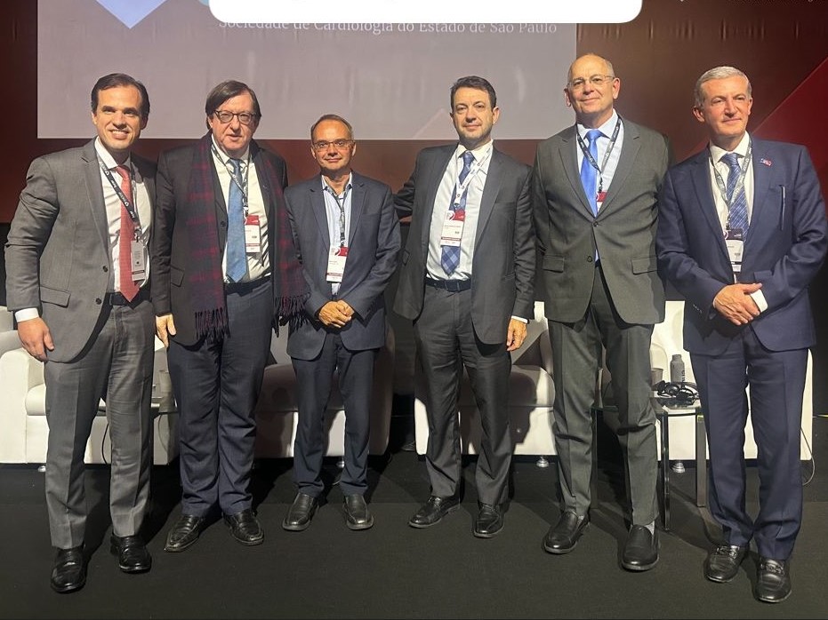 Seis homens posam juntos em pé sobre um palco durante um evento profissional. Todos vestem trajes sociais (terno e gravata) e usam crachás de identificação pendurados no pescoço. Ao fundo, vê-se parte de um telão com inscrições, parcialmente encobertas, onde se lê “Sociedade de Cardiologia do Estado de São Paulo”. O grupo está disposto lado a lado, sorrindo para a foto, e há poltronas brancas atrás deles, compondo o ambiente do auditório ou congresso.