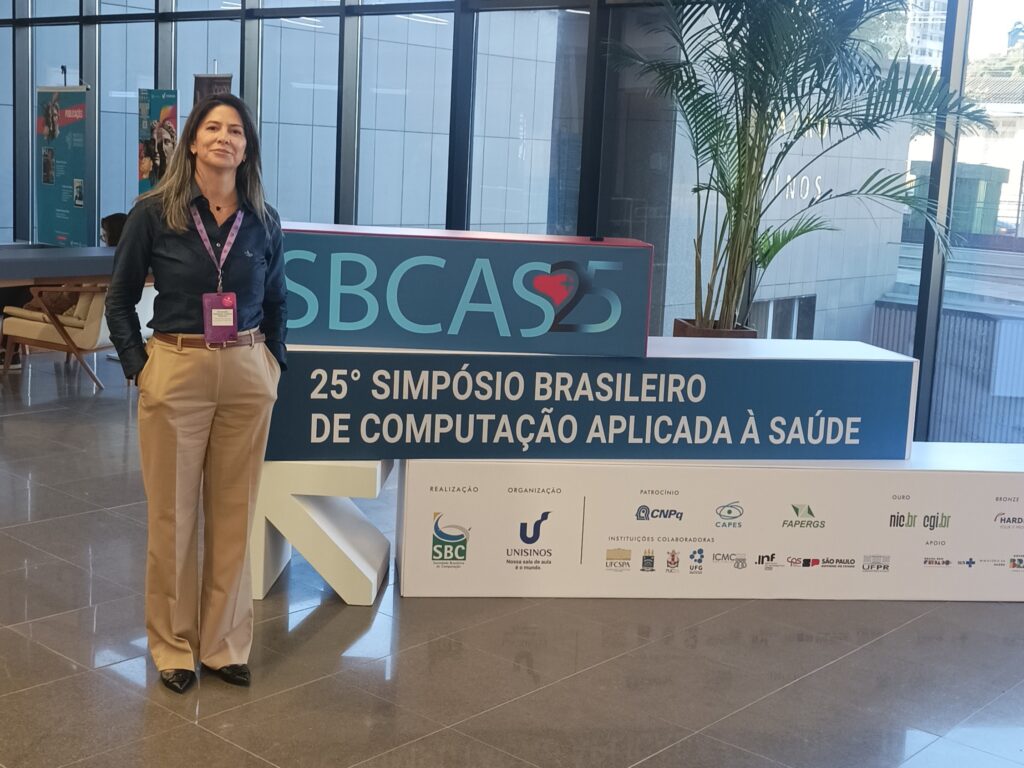Mulher de cabelos lisos e castanhos, usando camisa azul escura, calça bege e crachá roxo pendurado no pescoço, está em pé ao lado de uma estrutura com o logotipo “SBCAS 25” e a inscrição “25º Simpósio Brasileiro de Computação Aplicada à Saúde”. O ambiente é moderno, com piso de pedra polida, janelas de vidro amplas ao fundo e plantas decorativas. Ao fundo, há banners e móveis de recepção.