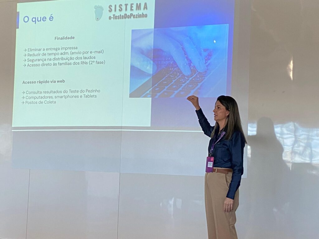 A mesma mulher está em frente a uma projeção na parede, apresentando um slide sobre o “Sistema e-TesteDoPezinho”. O slide mostra tópicos como eliminação de entrega impressa, redução de tempo administrativo e acesso via web a resultados do teste do pezinho. Ela está com o braço erguido apontando para a projeção. Seu reflexo aparece na parede brilhante ao lado.
