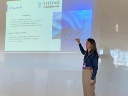 A mesma mulher está em frente a uma projeção na parede, apresentando um slide sobre o “Sistema e-TesteDoPezinho”. O slide mostra tópicos como eliminação de entrega impressa, redução de tempo administrativo e acesso via web a resultados do teste do pezinho. Ela está com o braço erguido apontando para a projeção. Seu reflexo aparece na parede brilhante ao lado.