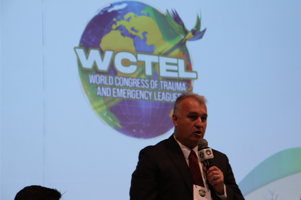 O Presidente do WCTEL (World Congress of Trauma and Emergency League), professor Gustavo Fraga, do Departamento de Cirurgia da FCM, posa ao lado do logotipo da organização. O logotipo exibe as letras 'WCTEL' em destaque, com o nome completo 'WORLD CONGRESS OF TRAUMA AND EMERGENCY LEAGUE' abaixo, em fonte moderna. O professor veste traje profissional, como paletó e gravata, e tem expressão serena e confiante, refletindo sua liderança na área de trauma e emergência. O fundo da imagem é neutro, destacando a figura do professor e o logotipo.