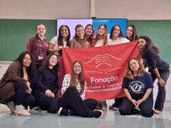 Um grupo de 13 pessoas posa sorridente em uma sala de aula, segurando uma bandeira vermelha com o logotipo e nome "Fonação – Assessoria e Consultoria em Projetos Fonoaudiológicos". Ao fundo, há um quadro verde e uma televisão.