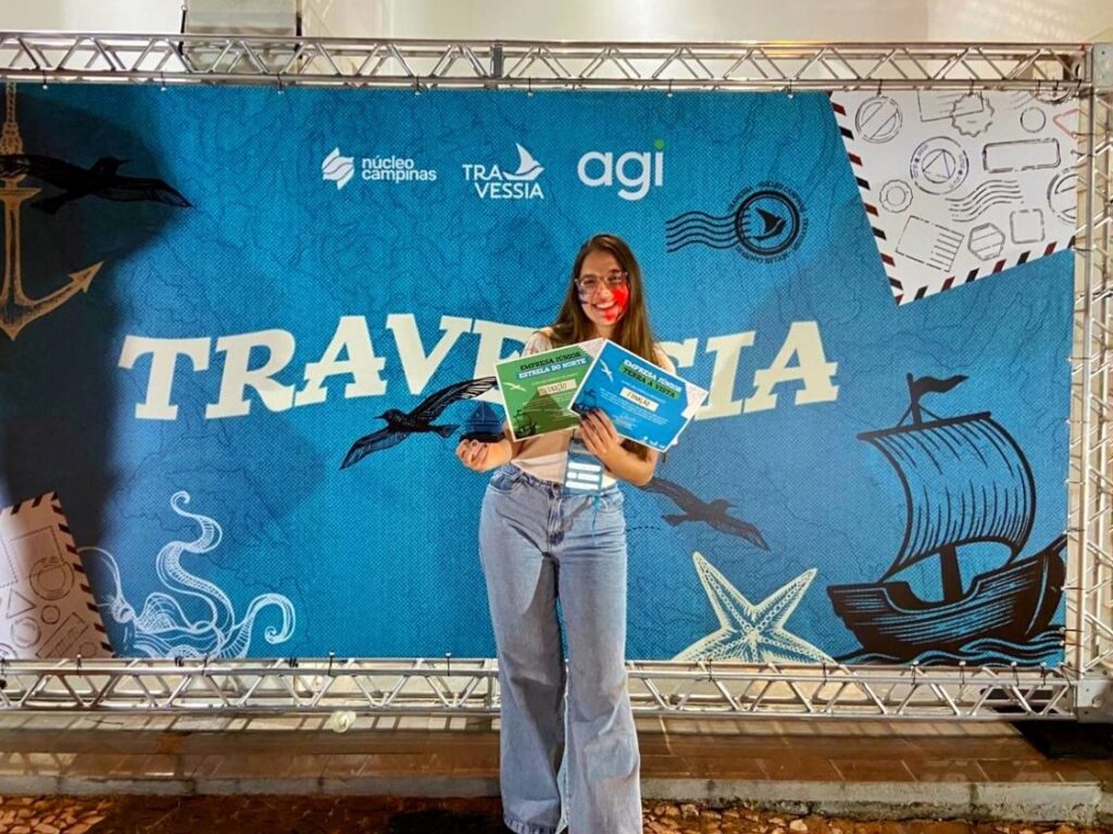 Jovem segura certificados e sorri diante de um painel azul decorado com elementos náuticos e a palavra "Travessia" em destaque. O fundo também exibe os logotipos do Núcleo Campinas, AGI e Travessia.