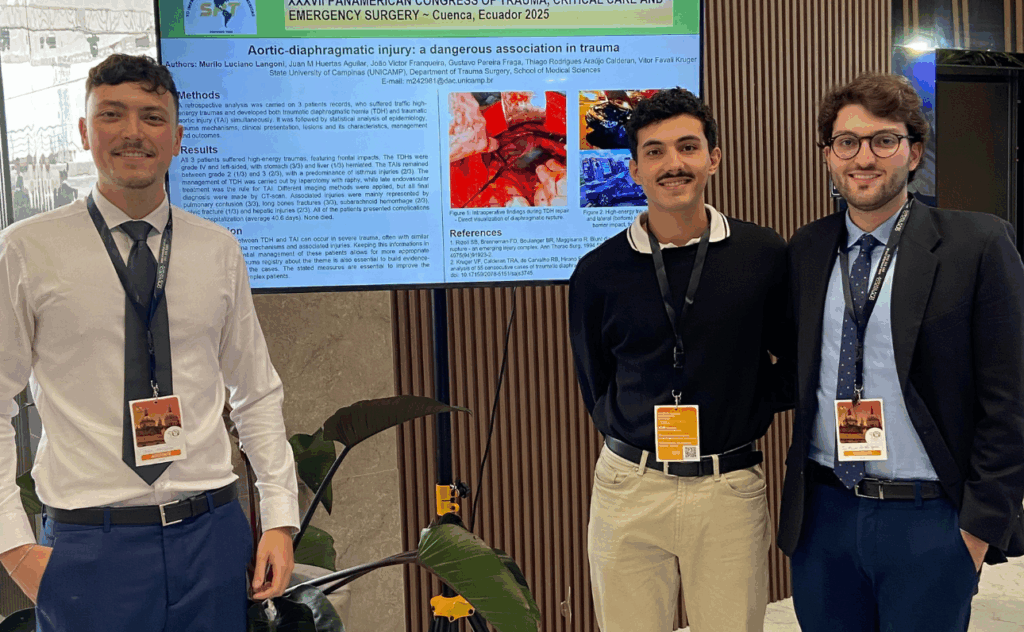 Dois homens estão posando para uma foto em um evento. À esquerda, um homem mais velho veste terno preto com camisa branca e gravata amarela, com um crachá pendurado no pescoço e um pequeno laço amarelo preso à lapela. À direita, um homem mais jovem, usando óculos e suéter preto, segura um certificado da "Sociedad Panamericana de Trauma" (Sociedade Panamericana de Trauma). Ambos estão sorrindo. Ao fundo, há um banner com imagens de prédios históricos e ruas de uma cidade, sugerindo um contexto acadêmico ou cultural.