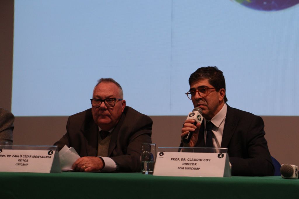O reitor da Unicamp, Paulo César Montagner, e o Diretor da FCM, Cláudio Saddy Rodrigues Coy, participam da mesa de abertura do WCTEL (World Congress of Trauma and Emergency League). À esquerda, o reitor veste traje social e tem postura solene, enquanto à direita, o diretor segura um microfone e está em momento de discurso. Ambos estão em um ambiente formal, possivelmente um auditório, com mesas de apoio e logomarcas do evento ao fundo. A imagem transmite a importância acadêmica e institucional do congresso.