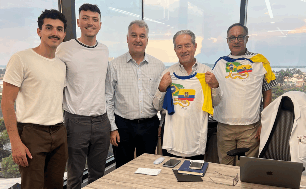 Cinco homens estão em uma sala com janelas amplas e vista para uma cidade ao fundo. Estão sorrindo e posando para a foto, próximos a uma mesa com objetos como celular, óculos, bloco de anotações e um notebook. Os dois homens à direita seguram camisetas brancas com detalhes em azul e amarelo, que exibem bandeiras do Brasil e do Equador, além do texto "Yellow Wave 2025". Os dois homens à esquerda usam roupas casuais (camisetas claras e calças escuras), enquanto os três à direita estão com camisas sociais. Uma cadeira ao lado exibe um jaleco branco com o nome “Dr. […] Cirugía”. O ambiente sugere uma reunião profissional ou preparativo para um evento futuro.